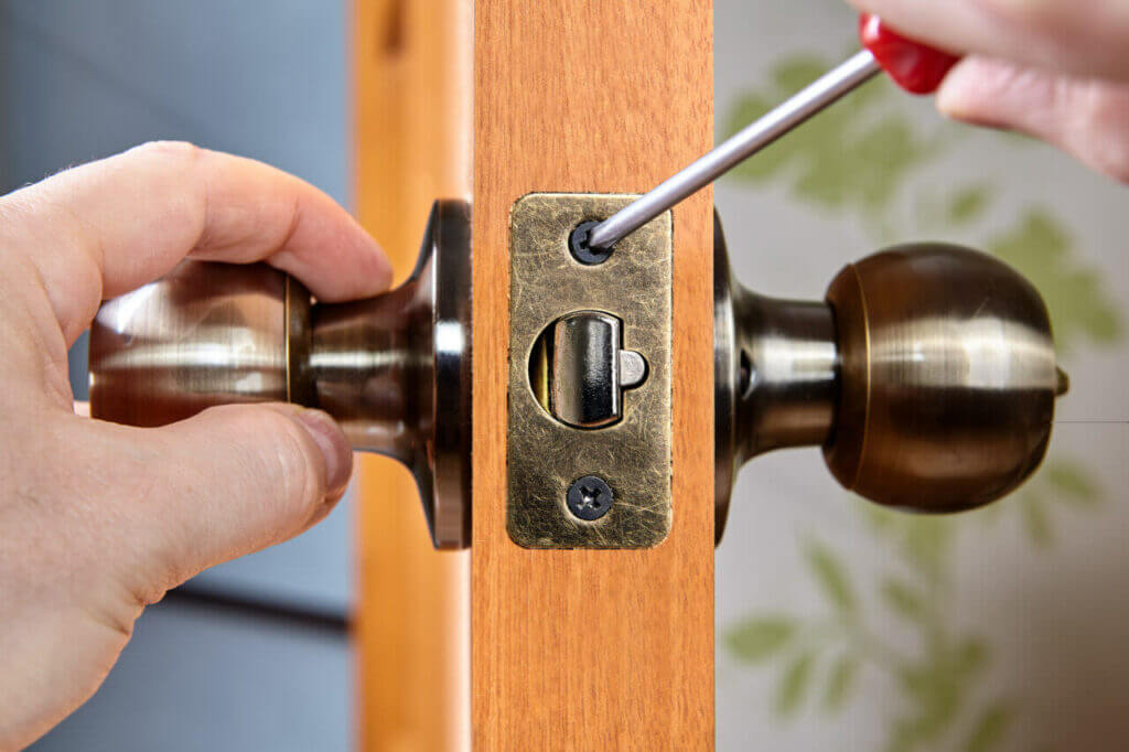 A locksmith is installing a brass door knob onto a wooden door using a red-handled screwdriver. One hand holds the knob's base plate while the other hand tightens the screws. The door is partially open.